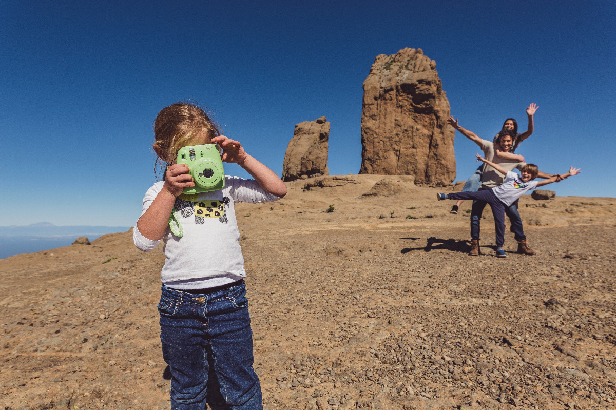 Mädchen fotografiert mit Sofortbildkamera vor Familie, die vor Roque Nublo auf felsigem Plateau posiert.
